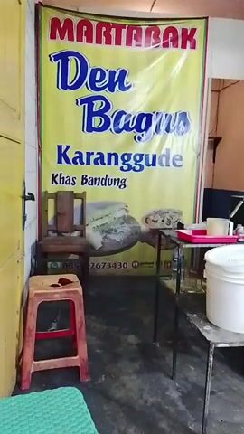 The video pans across a small food stall. A large yellow banner with red and blue text advertising "MARTABAK Den Bagus Karangdude Khas Bandung" is prominently displayed. To the left, a wooden chair and a red stool are visible. To the right, a metal table holds a white bucket, a red tray, and other containers. Further back, a gas stove and propane tank are present, along with cardboard boxes. A person in a blue t-shirt and blue jeans stands near a yellow door. The stall appears to be indoors, with tiled walls in the background. A menu board with various food items and prices is also visible. A QR code for payment is displayed on a stand.