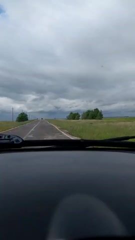 The video shows a first-person view from inside a car driving on a straight, empty road through a grassy, rural landscape. The sky is overcast with dark, heavy clouds. Trees are visible on either side of the road, and power poles line the left side. The car's windshield wipers are slightly visible at the bottom of the frame. The road appears to be wet, suggesting recent rain. The overall atmosphere is somewhat gloomy and isolated.
