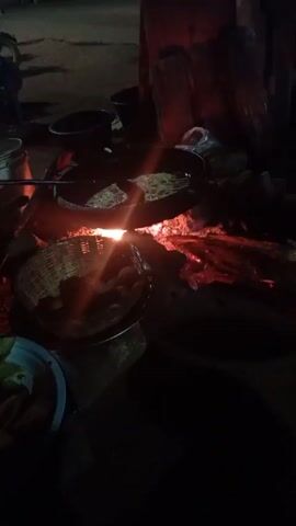 In a dimly lit outdoor scene at night, a person is cooking food over a wood-burning fire. Two pans are placed over the fire, with visible cooking taking place in each. A metal utensil is being used to stir or maneuver the food in one of the pans. To the left of the fire is a plate or bowl containing what appears to be previously cooked food. Other containers, possibly used for cooking ingredients or storage, are scattered nearby. The background suggests an outdoor or street-side setting, with faint lighting and the presence of vehicles and possibly structures in the distance. The focus of the scene is on the cooking process over the open fire, with the bright glow of the flames providing the main source of light.