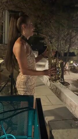 A woman plays a saxophone on a stone ledge at night. She is wearing a sparkly, sleeveless, one-shoulder dress. Her hair is pulled back in a high ponytail. Behind her, a large black speaker stands on a tripod. In the background, trees and outdoor seating with people are visible. The scene appears to be an outdoor restaurant or bar. The lighting is dim, suggesting it is nighttime.