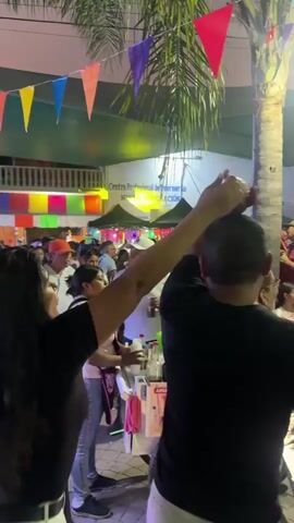 The video shows a nighttime scene in what appears to be a busy town square or outdoor market. Colorful triangular flags are strung overhead, and a few palm trees are visible. There are many people milling about, with food carts and stalls scattered throughout the area. A band is playing on a stage, lit with bright blue and red lights, and smoke effects are visible. People seem to be enjoying themselves, some dancing, others watching the performance.  The camera moves somewhat haphazardly through the crowd.