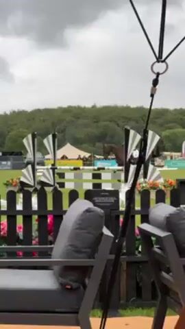Here's a concise description of the video:

The video captures a horse and rider, seemingly at an equestrian event, jumping over a fence decorated with black and white fan-like shapes and flowers. The horse is brown, and the rider is wearing a helmet and riding clothes. The video is shot from behind a partially visible outdoor lounge area with gray cushions. In the background, there's a grassy field, trees, flags, tents, and a few people near the fence. The sky is overcast.
