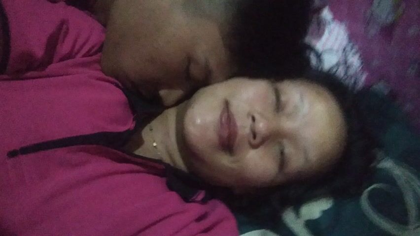 The video opens with a close-up of two individuals lying down, likely on a bed, engaged in an intimate moment. One person, with short dark hair, is positioned on the left, leaning into the head/neck area of the other. The second person, on the right, has dark hair and a bright, happy smile, revealing braces on her teeth. Both are wearing matching or similar pink tops with dark accents. A pink and white Hello Kitty patterned pillow is visible beneath them.

The camera then quickly pans away from the individuals, offering a brief glimpse of the room's interior. The setting appears to be a bedroom, characterized by a predominantly pink color scheme. Visible decorative elements include a pink mosquito net or canopy hanging from the ceiling, a pink dollhouse or shelving unit on the left, and green leafy garlands. The camera movement becomes increasingly shaky and blurred towards the end of the clip, suggesting low light conditions and unsteady handling.

The initial segment conveys a sense of happiness and affection between the two individuals. No discernible text is visible in the video. The location is identified as Kecamatan Cimaung, Indonesia.