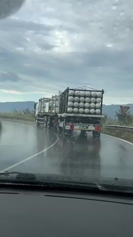 A dashcam view from inside a car shows a semi-trailer truck ahead on a wet, two-lane road. The truck is carrying numerous white, spherical tanks, likely gas cylinders, arranged in multiple rows and layers on its trailer. The road surface is visibly wet, indicating rain or recent precipitation, and raindrops are visible on the car's windshield. In the foreground, on the car's dashboard, a smiling banana-shaped plush toy with a pink flower clip on its head is visible. Mountains or hills can be seen in the distant background under an overcast sky.

Notable text on the back of the truck includes a number "1408Z" and the brand "КРОНЕ" (KRONE). Bulgarian license plates, "P 7273 TB" and "P 7273 TA," are also visible on the trailer. A hazard warning sign with "23" in a diamond and "19" in a rectangle can be seen on the back of the truck. The scene depicts travel on a potentially rainy day near Marino Pole, Bulgaria.