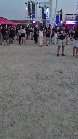 The video shows a crowded outdoor event, possibly a music festival, with large stage structures in the background, featuring bright lights and screens. A group of people are standing in the foreground, many looking at their phones. The ground is covered in gravel and dirt. The sky is a pale, dusky color, suggesting it is either early morning or late evening. The atmosphere appears relaxed as people mingle and wait.