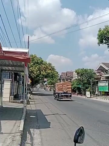 A street view on a sunny day. The road is paved and appears to be a two-lane road. On the left side of the road, there is a sidewalk with some buildings or shops. One building has a red awning. The sidewalk is shaded by overhead wires and possibly trees. On the right side of the road, there are more buildings, trees, and a few signs. A truck carrying what appears to be lumber or wood is driving away from the camera down the road. The sky is blue with scattered white clouds. The perspective suggests the video is being filmed from a moving vehicle, likely a motorcycle, as a portion of a mirror or handle is visible in the lower right corner of the frame.
