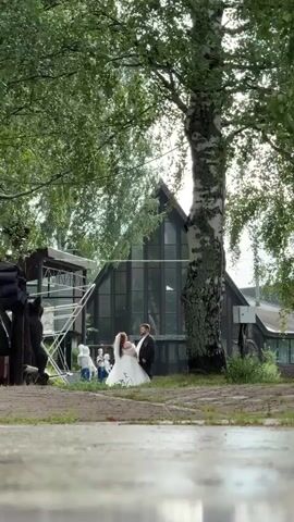 A wedding couple stands in front of a modern building with large windows. The bride wears a white wedding dress and veil, while the groom is in a dark suit. They are partially obscured by a large birch tree. To the left of the couple, an older man in a blue shirt and dark pants is visible, holding a dark jacket over his shoulder. Further left, there appears to be some kind of scaffolding or outdoor structure. In the foreground, a person's legs, clad in black pants and white sneakers, are visible, facing away from the camera and towards the wedding couple. The environment is outdoors with lush green trees and foliage. The ground in the foreground appears wet or reflective. The scene seems to be a wedding photoshoot or ceremony taking place outdoors near a building.