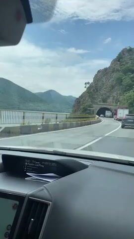The video, filmed from inside a moving car, shows a scenic drive in the Drobeta-Turnu Severin region of Romania. The car is traveling on a road that runs alongside a body of water, likely a river or lake, with lush green mountains in the distance and a rocky mountain face to the right. The sky is partly cloudy with patches of blue.

The vehicle approaches a stone-arched tunnel carved into the rock face. Several cars and a large truck are visible on the road ahead, with a black Volvo XC60 and a truck entering the tunnel as the camera gets closer. A "40" speed limit sign is visible next to the tunnel entrance. The car's dashboard is visible in the foreground, displaying "22.0 / 22C" and the word "Play," suggesting temperature readings and possibly a media player.