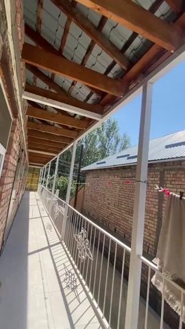 The video shows a long, covered walkway or balcony with a tiled floor and a white railing. The walkway is attached to a brick building. The roof of the walkway is made of wood beams and corrugated metal. The sky is blue and clear. There are some trees in the background and another building with a brick wall and a corrugated metal roof. A towel is hanging on a clothesline on the right side of the frame.