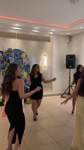 Several women in dresses dance in a room. The women, all in their twenties or thirties, wear black dresses and floral print. They dance in unison to music. A large speaker sits in the corner of the room, with a laptop in front of it.  The room has tiled floors, light walls, and decorative lighting along the ceiling. A floral arrangement is on the left side of the room.