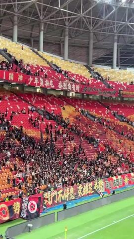 The video shows footage from inside a soccer stadium. In the first shot, the stands are partially filled with fans wearing mostly red and black colors, some holding up flags. There are banners along the bottom of the stands with Chinese writing on them. The second shot shows a crowd of people walking up stairs towards the soccer field. Many are wearing red shirts. The third shot shows the soccer field from the stands, and the final shot shows soccer players walking around on the field. There are large red banners with Chinese writing on them covering part of the stands.