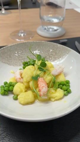 The video captures an indoor scene in what appears to be a modern dining or kitchen area. The setting features a combination of light wood and white tabletops, creating a clean aesthetic.

In the foreground, on a wooden table, a white plate holds a serving of gnocchi, green peas, shrimp, and microgreens, indicative of a prepared dish. Alongside the plate, there's a wine glass containing white wine and a water glass resting on a white coaster. A silver knife is placed on a black placemat, next to a black folder or menu.

The camera pans to an adjacent white tabletop where several raw salmon fillets are neatly arranged on a black grill pan or serving tray. Two glass water bottles are also visible on this table. A white card with "ONYX" printed on it is placed near the bottles.

A person wearing yellow clothing is briefly visible in the background as the camera moves. The overall impression is a culinary event, a cooking demonstration, or a meal being served, possibly in a professional or test kitchen environment. The location context for this media file is Rho, Italy.