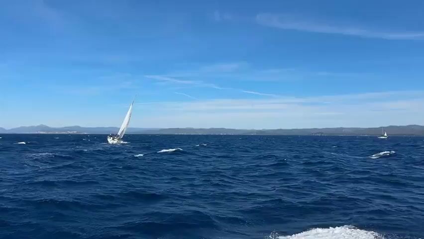 The video shows a sailboat with a white sail cutting through choppy blue water under a bright blue sky with wispy clouds. The sailboat is leaning to its side, indicating it is moving quickly. In the background, a distant coastline is visible with hills and some buildings. Another sailboat is visible further in the distance. The waves are creating whitecaps as the sailboat moves through them.