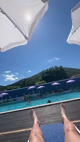 The video shows a person's legs and feet resting on a blue mat on a wooden deck. Two large white umbrellas are visible in the upper part of the frame, creating shade. The camera is pointed upwards, towards a bright blue sky with some clouds. In the background, there is a swimming pool with several people in it. The pool is surrounded by a blue wall, and purple umbrellas are set up along the poolside. Beyond the pool, there are green hills covered with trees. The overall scene depicts a sunny day at a swimming pool.