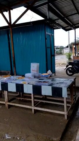 Under a corrugated metal roof supported by wooden beams, a roadside food stall is in operation. A long wooden table, covered with a black and white checkered cloth, displays numerous plastic containers filled with food items. Several people are gathered around the table, browsing and selecting items. Two men are standing behind the table, presumably serving customers. A red truck and a blue truck pass by on the road in the background. The ground appears damp and possibly muddy. A blue corrugated metal structure stands to the side of the food stall. A motorcycle is parked nearby.