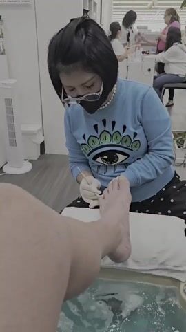 A person, from the knees down, is having their foot placed in a foot spa. A nail technician, wearing a blue sweatshirt with an eye design, a pearl necklace, glasses, and a face mask, is seated and using tools to work on the person's toenails. The foot spa is filled with bubbling water. In the background, other people are visible in what appears to be a nail salon.