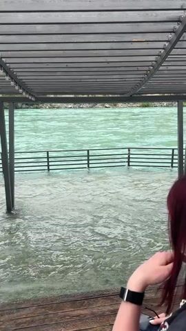 A woman with reddish-brown hair, wearing a dark t-shirt and carrying a red tote bag, stands on a partially submerged wooden deck under a slatted wooden pergola. She is observing the high water levels of a wide, turquoise-colored river. A dark metal fence is also partially submerged in the churning water. The woman initially looks towards the water, briefly turns her head towards the camera with a neutral expression, then turns back to face the river as she walks slightly forward. The scene appears to be Miatli, Russia, during daytime, possibly with overcast weather given the diffused light, highlighting the unusually high river level.