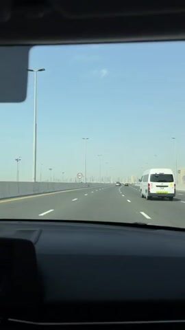 The view from inside a car driving on a multi-lane highway. The highway is mostly straight and relatively empty, with only a few other cars visible. The speed limit is 80. The city skyline is visible in the distance, with many tall buildings. The sky is clear and blue. The car's dashboard is visible in the foreground.