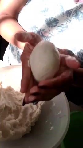 A person forms a ball of dough with their hands. They wear a white tank top with a floral pattern and have dark nail polish on their nails. A white bowl containing more dough is visible to the left. The background is out of focus.