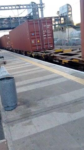 A freight train with multiple red shipping containers moves past a train station platform. The train is traveling from right to left across the frame. On one of the containers, the text "ZIM" is visible, while another container displays "The" followed by a colorful, stylized logo. The platform features concrete paving with a yellow tactile strip along its edge. In the foreground, a gray, perforated metal trash can is positioned on the platform. In the background, beyond the train tracks, there are fences, several parked cars, and an elevated structure, possibly an overpass or part of the station infrastructure. The sky is bright, suggesting it is daytime.