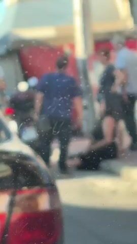 The video shows a street scene in Peristeri, Greece, during the daytime, likely late afternoon given the warm lighting. In the foreground, the rear of a dark-colored car is visible. The main focus is on a group of people on the sidewalk. A woman in a black dress is sitting on the curb, holding a phone and gesturing with her hands, appearing to be in conversation or distress. A man in a blue shirt and black pants stands near her, facing away from the camera, also with a bag slung across his body. Behind them, an older man in a light blue shirt and an older woman in a dark patterned dress stand together, observing the scene. A motorcycle with license plate "NYK 624" is parked nearby. The background shows a building with red and white elements, possibly a shop or restaurant, and graffiti on a wall. The overall atmosphere seems tense, with people gathered around the seated woman.