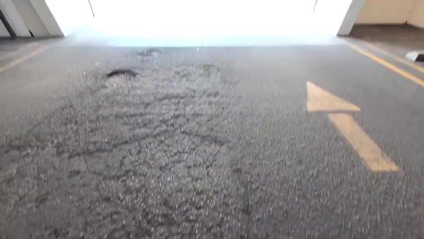 The video shows a view from inside a parking garage, looking out towards the entrance. The ground in the foreground is asphalt with several potholes and an arrow painted on it. Beyond that, there is a concrete area with lines painted on it, and a sign that reads "PARKING ENTRY" in reverse. There are caution signs visible above the entrance. Outside the garage, there is a parking lot with some trees and a building in the background. The lighting suggests it is daytime.