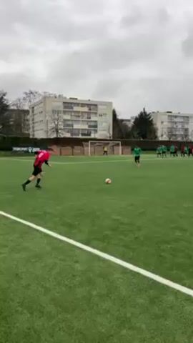 The video shows a soccer field with players practicing a free kick. A player in a red and black jersey is preparing to kick the ball towards the goal. In front of the goal, several players in green jerseys form a defensive wall. The goalkeeper stands ready to defend the goal. There are buildings visible in the background, and the sky is overcast. The video appears to be taken at ground level, providing a clear view of the field and the players.