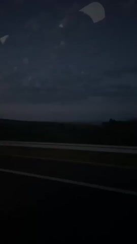 The video depicts a view from inside a vehicle moving along a highway at night or in very dim light. The sky above is dark blue, showing a few light clouds. The horizon line is visible, with a darker band of land or trees. The highway features concrete barriers with painted white lines marking the lanes. The vehicle is moving at a steady pace, and the focus is on the outside view. There is a fleeting blur of a passing vehicle in one frame.