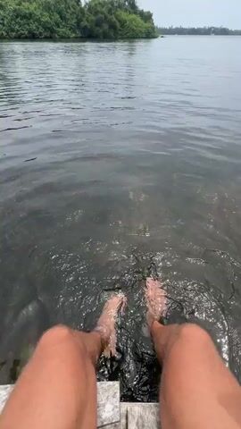 The video shows a first-person perspective of a person sitting on the edge of a wooden dock with their legs and feet in the water. The water is dark and murky with ripples and splashes created by the movement of the person's feet. In the background, there is a body of water stretching out towards a lush green island with palm trees. The sky is overcast. The scene appears to be a calm and natural environment.