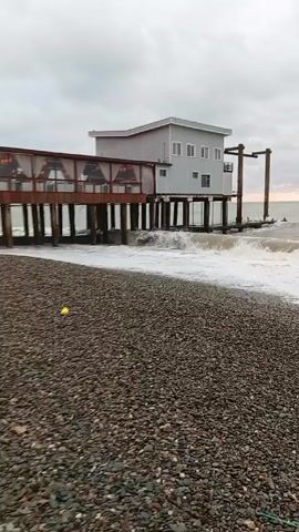 The video shows a pebble beach with a building on stilts extending into the ocean. The building has a covered walkway with tables and a taller section with windows. Waves crash against the stilts. There is a yellow object on the beach. The sky is overcast. The waves are coming closer to the beach.