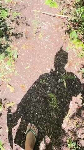 The video begins with a close-up of a foot wearing a flip-flop stepping onto a dirt path in a sun-dappled wooded area. The camera then pans up and to the right, showing a densely vegetated area with green foliage and scattered leaves on the ground. The scene shifts again to a wider view, revealing a stone pathway leading towards a swimming pool and a large white house. The pathway is bordered by manicured hedges and lush green plants. Beyond the pool, a patio area with wooden chairs and a large umbrella is visible. The video concludes with a shot of the clear blue water of the swimming pool reflecting the bright sky and surrounding trees.