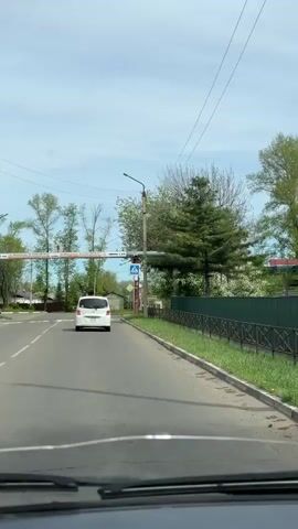 The video shows a vehicle's perspective driving down a paved road on a sunny day. A white minivan drives ahead. Overhead signs indicate street names and distances. There's a marked crosswalk with yellow stripes ahead, and pedestrian crossing signs are visible. Trees and bushes line the road, and houses are visible in the background. Power lines crisscross above the road. The sky is mostly clear with some wispy clouds.