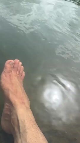 The video opens with a close-up of a person's bare foot resting in shallow water, with the foot partially submerged. The water is murky and reflects the cloudy sky. The camera then pans out to reveal a body of water, possibly a river or lake, with a rocky shore and a lush green hillside covered in trees. The sky is overcast, suggesting it might be a cloudy or rainy day. The overall scene depicts a natural, outdoor environment.