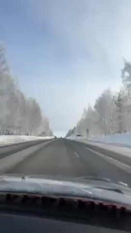 The video shows the view from inside a car driving down a road. The road is straight and has two lanes going in each direction, separated by a median. The road surface is dark and appears to be clear of snow and ice.  On both sides of the road, there are trees covered in frost or snow. The sky is light blue and slightly overcast. There are a few cars visible in the distance driving in the same direction.  There is a speed limit sign on the right side of the road and another sign further down. The dashboard of the car is visible at the bottom of the frame.