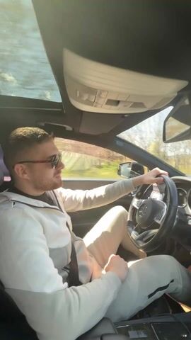 A man is driving a Mercedes-Benz. The camera is mounted on the ceiling of the car, providing a clear view of the driver, the dashboard, and the road ahead. The man is wearing sunglasses and a light-colored sweatsuit. The car has a sunroof, and the interior is gray and black. A VIP-branded air freshener hangs from the rearview mirror. The driver's left hand is on the steering wheel. The weather is sunny, and the road appears to be winding through a rural area. Music is playing.