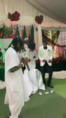 Sure, here is a description of the video:

The video shows a wedding reception. The bride and groom, in white outfits, are dancing with a guest in a white robe. The event appears to be indoors, with a tent setup and decorations. Money is scattered on the ground near the couple. The guest in the robe dances and sings into a microphone.