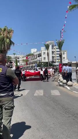A group of people, including young students, are participating in a parade. They are carrying a very large red flag with white crescent and star, the national flag of Turkey. The students are dressed in school uniforms, consisting of white shirts and tan skirts. Some adults are also present, with one man in a black shirt and pants walking alongside the students. The parade is taking place on a street with buildings and palm trees lining the sides. Turkish flags and red and white bunting are visible decorating the buildings. The sky is clear and blue. The camera is focused on the parade participants as they walk across a pedestrian crossing.