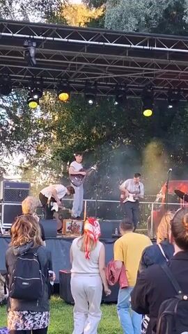 A band performs on an outdoor stage under a truss structure with lights. Two guitarists are on stage, one in the center playing a white guitar and another to the right playing a red guitar. A person in the background bends over on the stage. A crowd watches from the foreground, with trees and foliage visible behind the stage. The sky appears bright, suggesting daylight or early evening. One person in the crowd wears a blue hooded jacket and a black baseball cap, facing the stage. Another person in the crowd wears a red bandana. The overall atmosphere appears to be a live music event.
