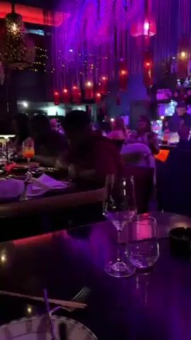 The video shows people eating at a restaurant, bar, or club. The lighting is primarily purple and red. There are two people seated next to each other, with one man sitting closest to the camera wearing a maroon shirt and several gold bracelets. He is looking down at his hands. On the table in front of him are sunglasses, a white napkin, and what looks like a plate. A wine glass and a shorter drinking glass are in front of the camera. The room is decorated with red light fixtures hanging from the ceiling.