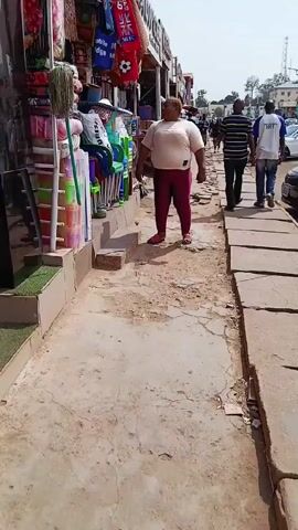 The video shows a woman standing on a sidewalk next to a shop. She is wearing a light-colored shirt and dark leggings. The shop is selling a variety of goods, including furniture, bags, and other household items. The sidewalk is cracked and dusty. There are other people walking on the sidewalk in the background. The video is shot from a handheld camera.