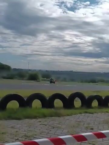 A person on a white and green motorcycle drives on a gray paved racetrack on a cloudy day. The bike is moving left-to-right, away from the camera, in the center of the shot, behind a row of black tires. The landscape beyond the racetrack includes trees and low-lying hills. The camera follows the motorcycle briefly before stopping.