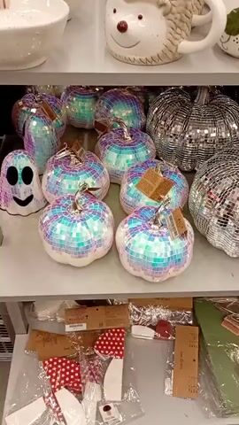 The video shows a display of Halloween decorations on shelves. The items prominently featured are disco ball-like pumpkins and a ghost. The pumpkins are white with mosaic tiles in iridescent shades of pink, blue, and green. The ghost is also covered in similar mosaic tiles and has black eyes and a black smiling mouth. The decorations are arranged on what appears to be retail shelving. Below the Halloween decorations, there are various other packaged items on a lower shelf, including what look like holiday-themed fabric items (e.g., red and white polka dot fabric) and tools or craft supplies. The lighting in the scene is bright, suggesting an indoor retail environment. There are no people visible in the video.
