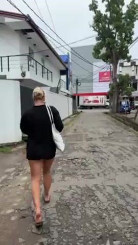 A woman with blonde hair is walking away from the camera down a potholed road. She is wearing a black jacket, shorts, and sandals, and carrying a white tote bag. On the left is a white building and on the right is a tree. Above, there is a tangle of power lines. A motorcycle passes her on the road. Further down the road there is a truck with a red and white sign. The scene is overcast.