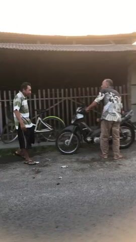 The video shows two men standing outside next to a motorcycle and a bicycle. The man on the left is wearing a striped shirt and shorts, and is facing the man on the right. The man on the right is wearing a patterned shirt and pants, and is facing the man on the left. The men appear to be talking to each other. A fence is visible behind them, and a building is in the background. The lighting suggests it is either dawn or dusk.