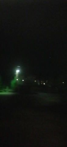The video shows a dark scene at night.  A streetlight with a green hue is visible in the distance, and there are a few other blurry lights further away, possibly from houses. The camera pans slightly to the right, showing more of the dark environment before briefly pointing upwards towards a light source. The scene is blurry and lacks detail, emphasizing the darkness.