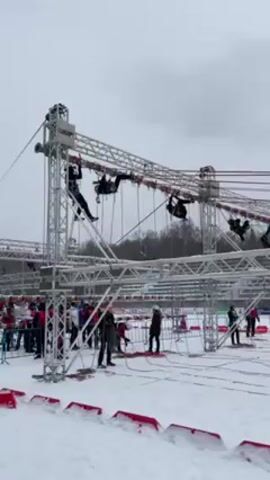 Several people are navigating a complex obstacle course in a snow-covered outdoor environment. The course is constructed from a network of interconnected metal structures and ropes. Participants in black attire are seen hanging and traversing various sections of the course, suspended in the air. Below them, on the snowy ground, a crowd of spectators, dressed in a mix of red and dark-colored clothing, are observing the event. Additionally, some individuals in dark jackets and casual clothing are seen adjusting ropes and equipment around the base of the structure. The sky above is overcast, indicating cold weather conditions.