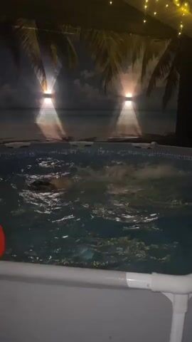 A young girl is swimming in an above-ground pool at night. The pool is filled with rippling, clear water. The girl is wearing a blue bikini top and has her hair pulled back. She splashes water on her face and raises her arms in the air. An orange pool noodle is visible on the side of the pool. The backdrop behind the pool is a mural of a tropical beach with palm trees, lit by two sconces. String lights are visible in the top of the frame. The pool is surrounded by a white frame.