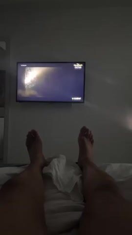 A person lies in bed with their feet pointed towards a television mounted on the wall. The television is displaying a scene from a program. The room is dimly lit. A white cloth is resting on the bed between the person's legs. The bed appears to be made with white linens. The wall is a muted gray color. The person's legs are visible from the upper thigh down.