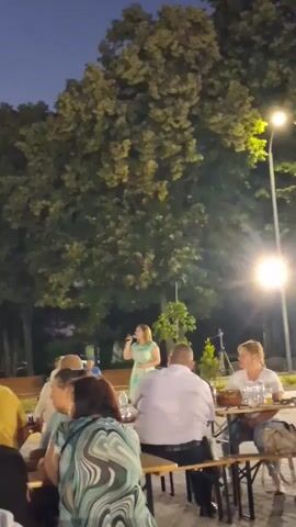 The video shows an outdoor evening gathering with a woman singing into a microphone on a small stage. Several people are seated at picnic tables in front of the stage, some facing the singer and others with their backs to the camera. A large tree with dense foliage dominates the background, and a streetlamp with a bright light is visible to the right. The sky is dark blue, indicating it is dusk or early night. The singer is wearing a light-colored dress. Some of the seated individuals are also facing the singer and appear to be listening. The overall scene suggests a casual outdoor event, possibly a concert or performance.