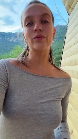 A woman with her hair in a ponytail, wearing a grey off-the-shoulder top and grey sweatpants, poses for a video. She is standing next to a wooden structure, with a mountainous landscape and greenery in the background. The sky is blue with some clouds, suggesting it is daytime. The woman smiles and playfully poses, at one point touching her hair and then turning to the camera with a wide smile. She has earrings and a watch on her left wrist. The setting appears to be outdoors, possibly in a rural or mountainous region, consistent with Bistrica, Montenegro. The overall mood is lighthearted and happy. There is no visible text in the video.